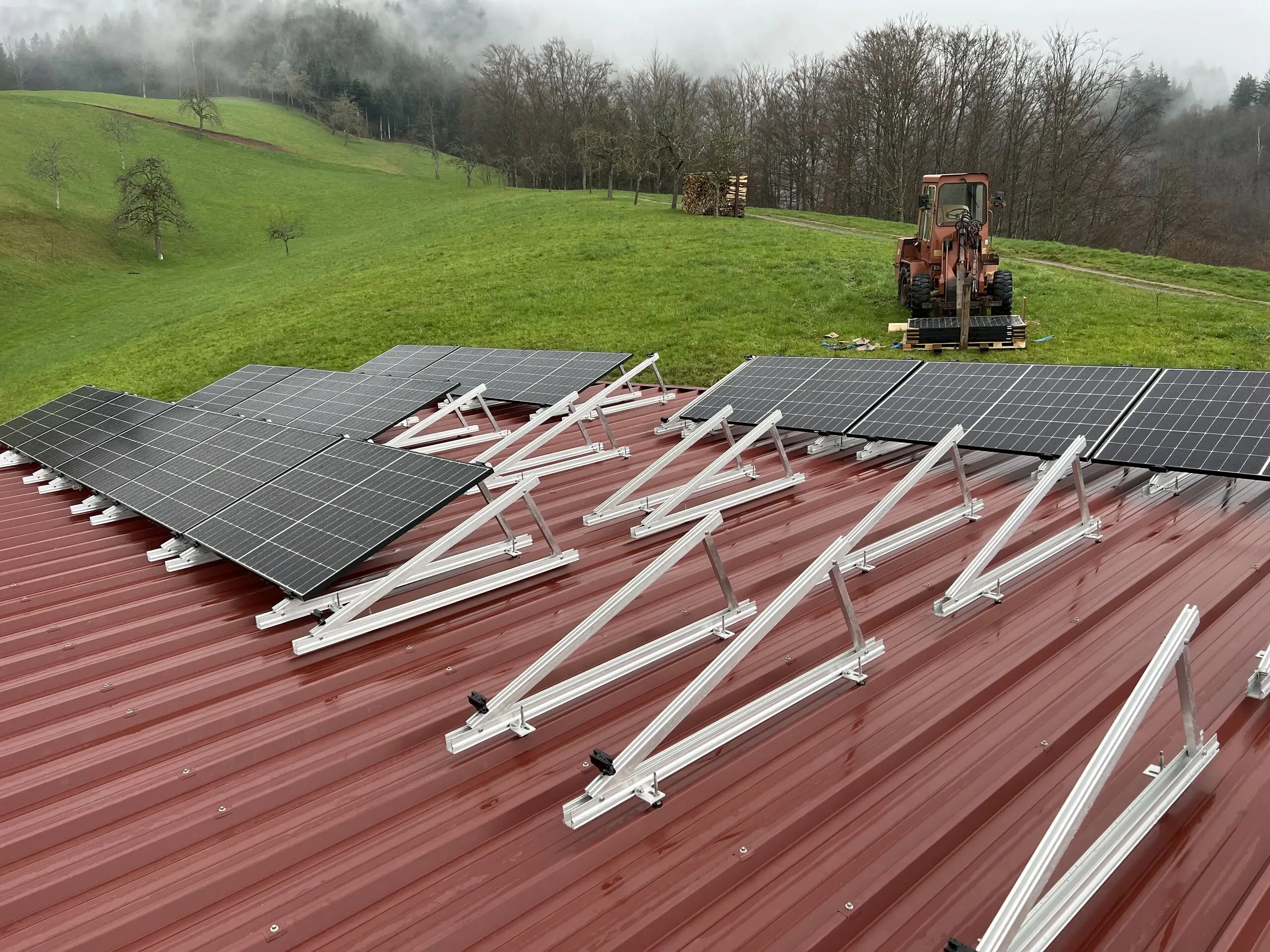 Photovoltaikmodule in einer aufgeständerten Unterkonstruktion aus Aluminium auf einem roten Trapezblechdach, ein Teil der Gestelle ist noch unbelegt, im Hintergrund eine Wiese mit einem Radlader