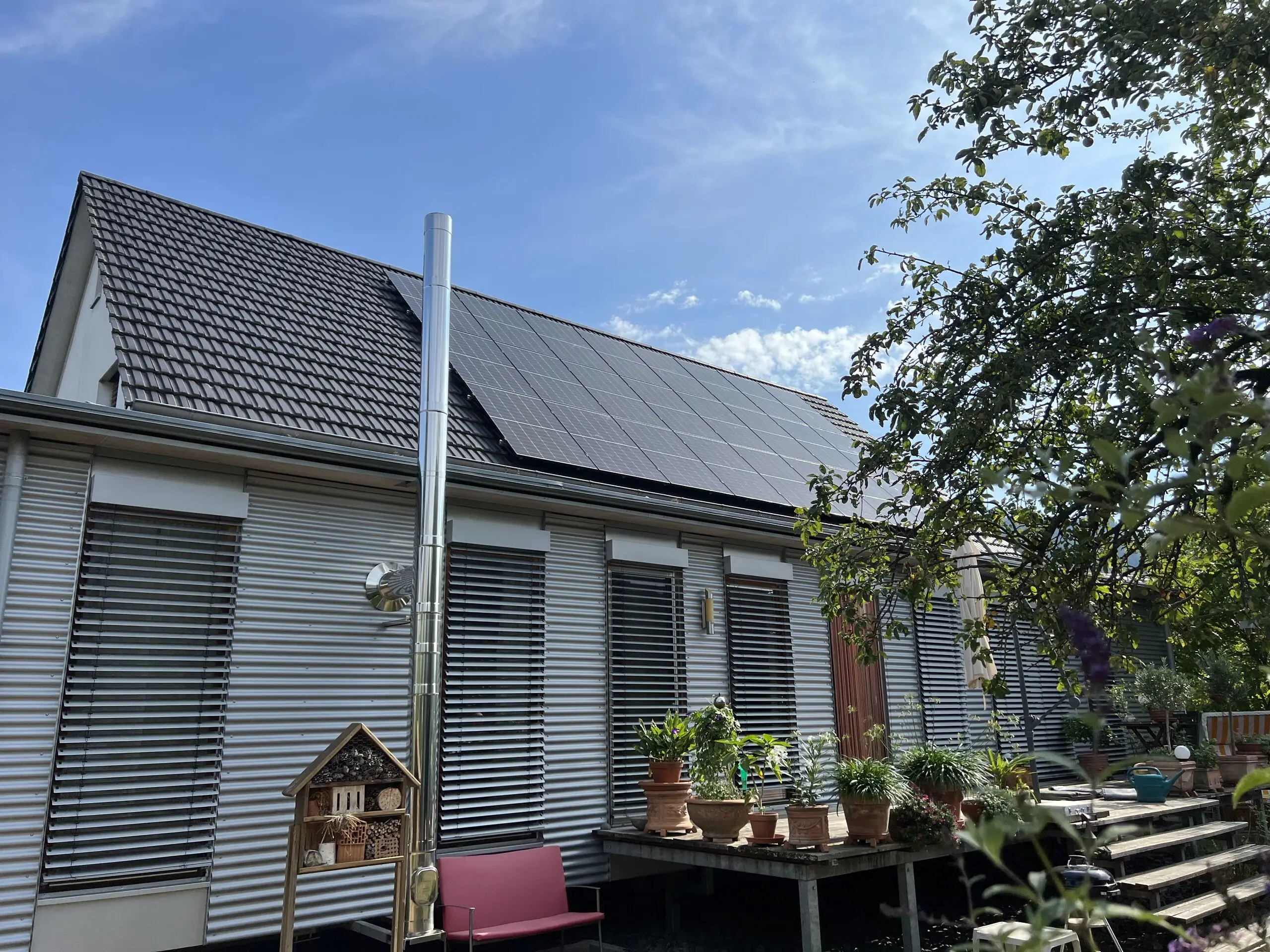 Wohnhaus mit Metallfassade und schwarzen Photovoltaikmodulen auf dem Satteldach, davor eine Terrasse mit Pflanzen