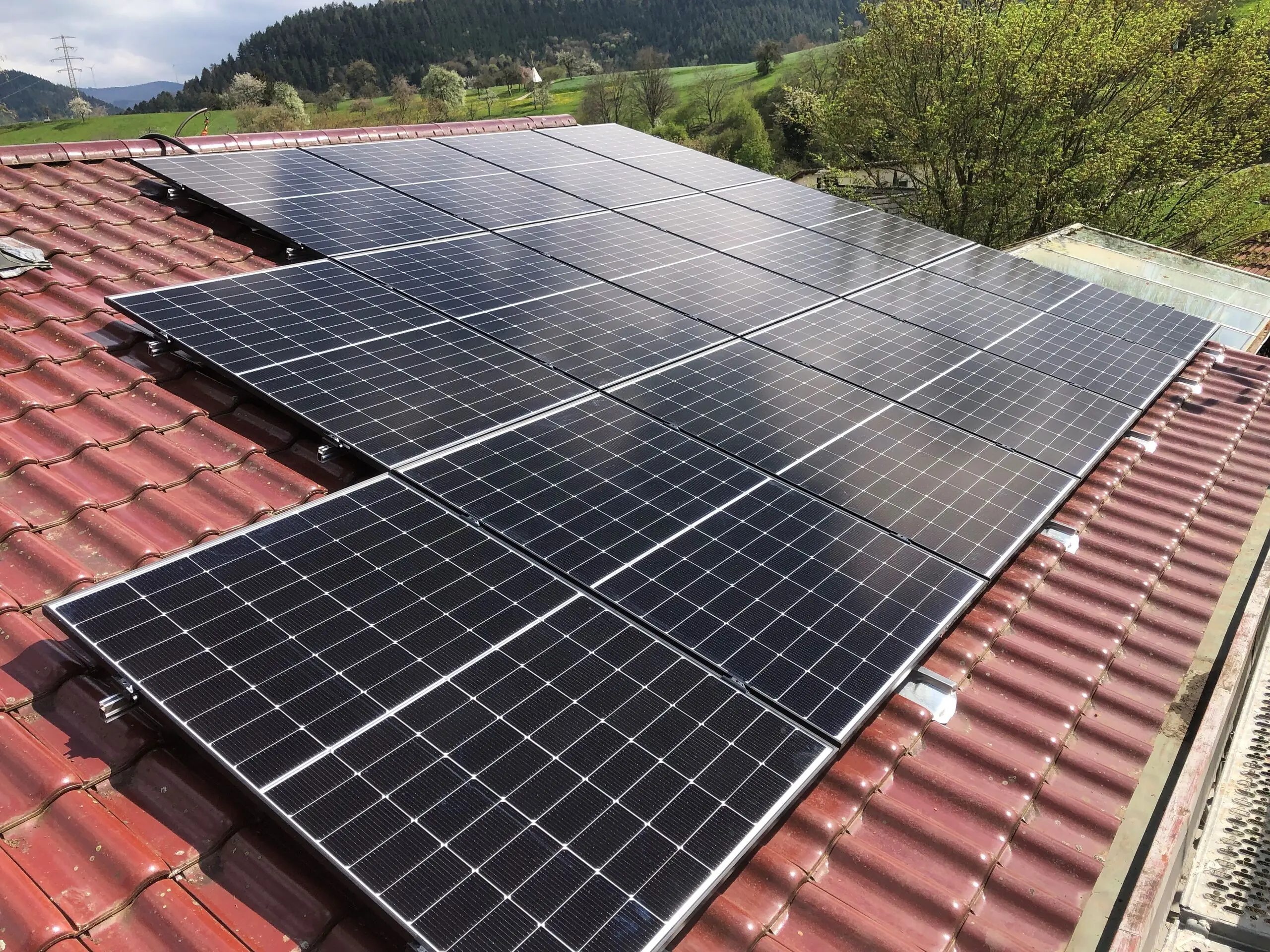 Photovoltaikmodule auf einem roten Ziegeldach, dahinter blühende Obstwiesen und bewaldete Hügel