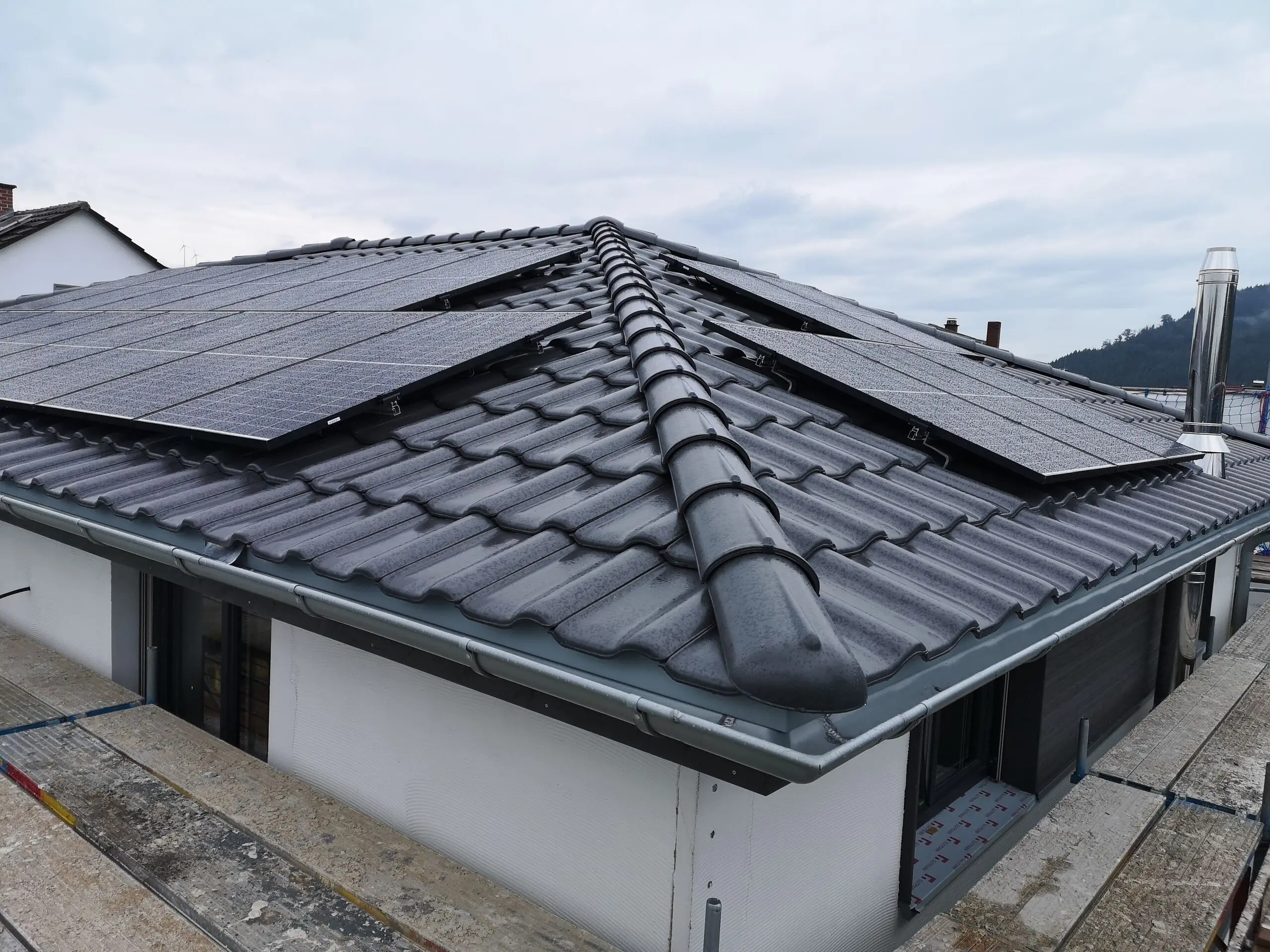 Anthrazitfarbenes Walmdach eines Bungalows, darauf Photovoltaikmodule in zwei Reihen, im Hintergrund bewaldete Hügel